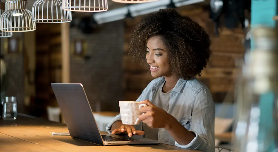 Image of a woman working on a laptop computer and holding a cup of coffee.