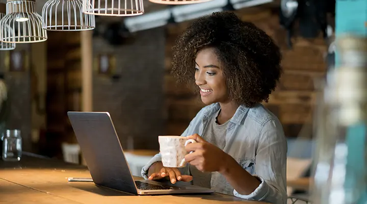 Image of a woman working on a laptop computer and holding a cup of coffee.
