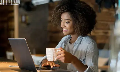 Image of a woman working on a laptop computer and holding a cup of coffee.