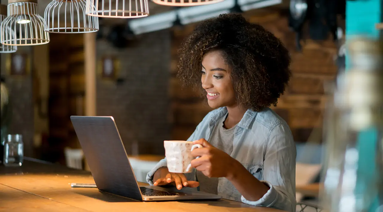 Image of a woman working on a laptop computer and holding a cup of coffee.